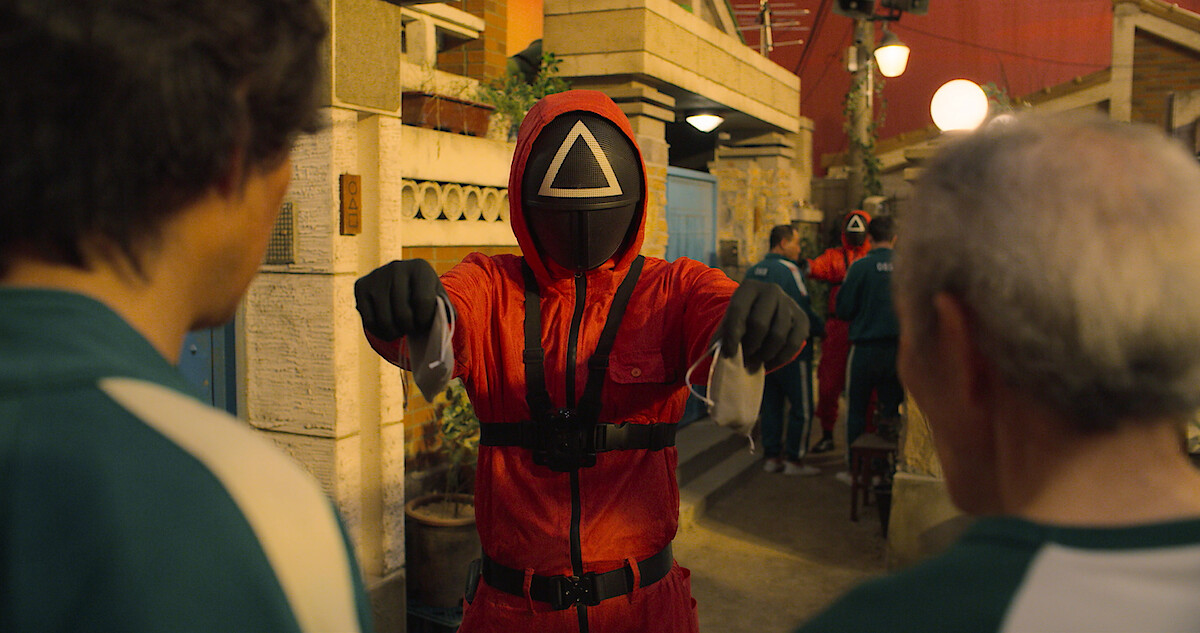 Person wearing red jumpsuit and a black mask with a white outlined triangle, covering their face.