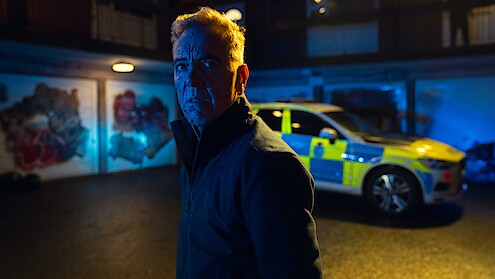 A man with blue markings on his face stands in a dimly lit urban parking area at night, with a police car and graffiti-covered garage doors in the background, illuminated by blue and yellow lighting.