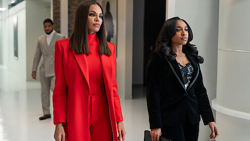 Crystle Stewart as Mallory and Taylor Polidore Williams as Kimmie in business attire, one in a red suit and the other in a black suit, walk confidently down a modern office hallway, with a man in a grey suit walking behind them.