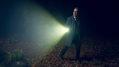 Man in suit and overcoat searching a dark, foggy forest at night with a flashlight, fallen leaves and debris around, tense and mysterious atmosphere.