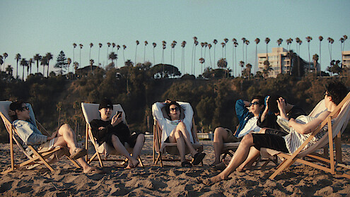 j-hope, Suga, Jin, RM, Jung Kook, and Jimin relaxing on beach chairs in the sand, enjoying a sunny day. Palm trees and buildings are visible in the background.