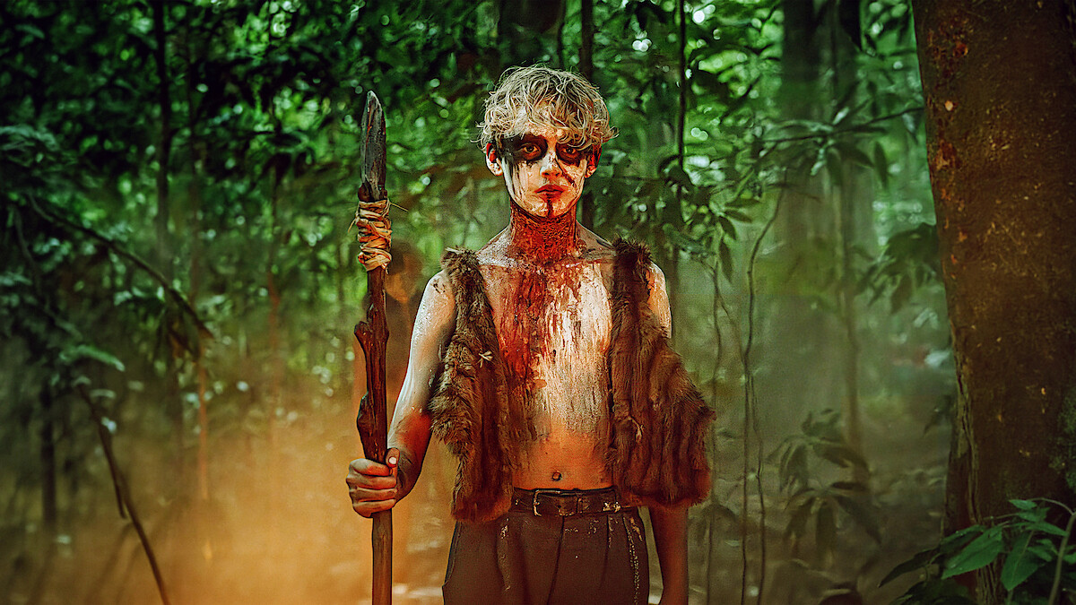 A person with painted face and fur vest stands holding a spear in a dense, misty, green forest, suggesting a primitive or survival setting.