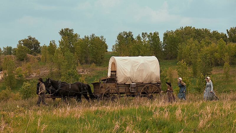 A covered wagon pulled by horses travels through a grassy field with three women and a child walking beside it, surrounded by trees under a partly cloudy sky.