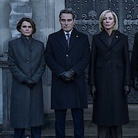 Three people in dark formal coats and gloves stand outside in front of an old stone building and ornate doors, posing seriously for a photograph in a somber or official setting.