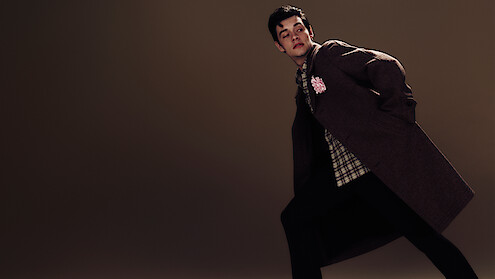 Louis Partridge stands on a dark brown background leaning over in an angled manner waring a brown coat with a plaid shirt and dark trousers.
