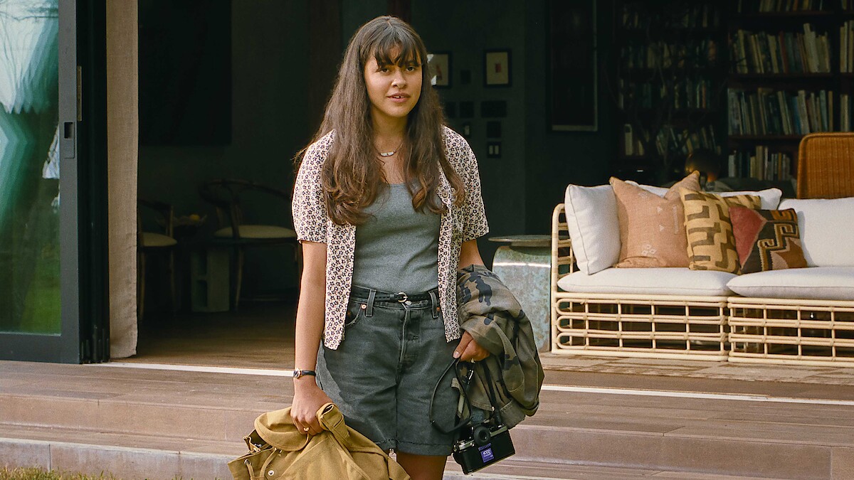 Young woman with long hair stands outside a cozy, modern house holding a coat, bag, and camera. The relaxed setting features a wicker sofa and bookshelves, evoking a calm, casual, and welcoming atmosphere.