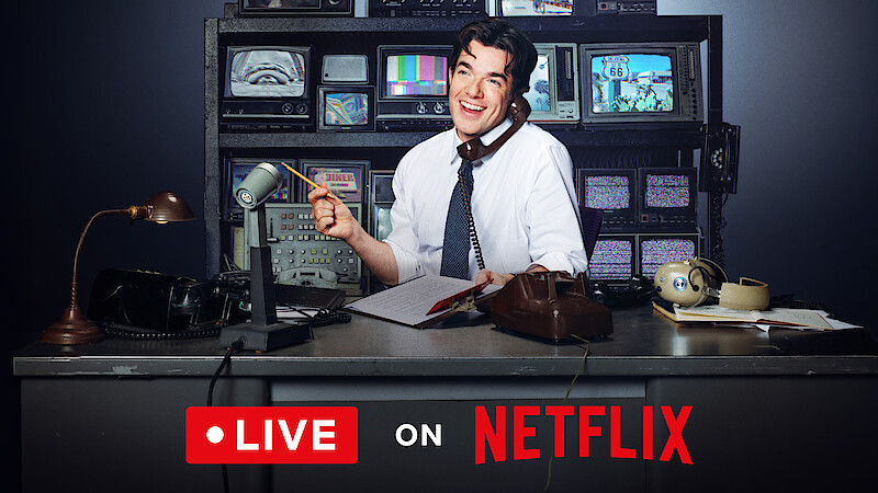 John Mulaney sitting at a desk in front of a wall of TVs on the phone.