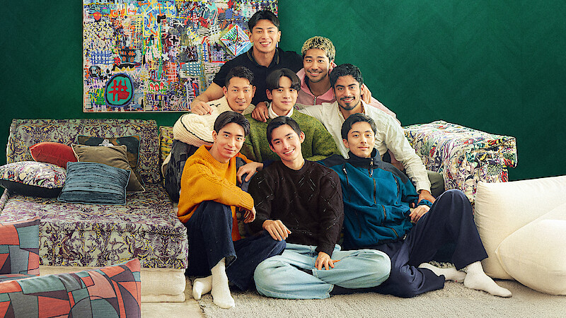 The cast of 'The Boyfriend' Season 2 sit closely together on a colorful sofa in a cozy living room with eclectic pillows, patterned furniture, and a vibrant art piece on a green wall, smiling and posing for the photo.