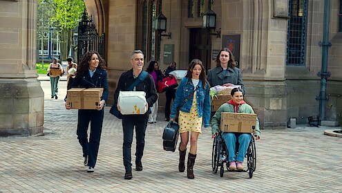 Five people carrying boxes and bags, including one in a wheelchair, walk together outside a historic stone building with arches and lanterns, suggesting a college or university move-in day.