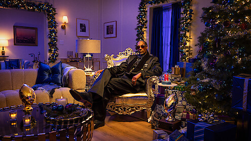Man in a suit sits on a luxurious chair in an elegant, festively decorated living room with Christmas tree, gifts, candles, and warm lighting, creating a cozy and celebratory mood.