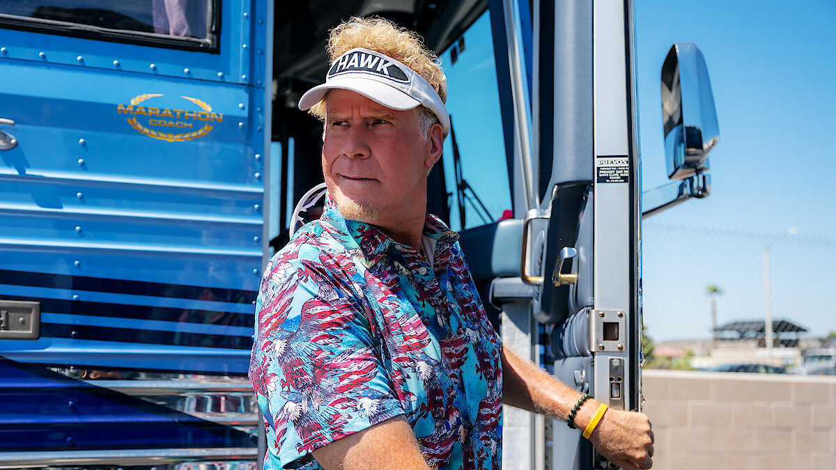 Man in a tropical shirt and visor stands at the doorway of a blue luxury bus on a sunny day, holding the door handle, with a fence and clear sky in the background.