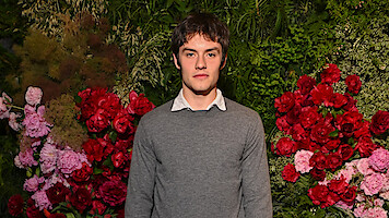 Young man in a gray sweater stands in front of a vibrant floral wall with red and pink flowers and green foliage, creating a natural, garden-like backdrop. Indoor setting, formal or event atmosphere.