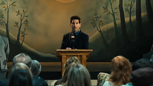 Man in a suit speaking at a wooden podium on stage with a microphone, addressing an audience in a room with a painted forest backdrop.