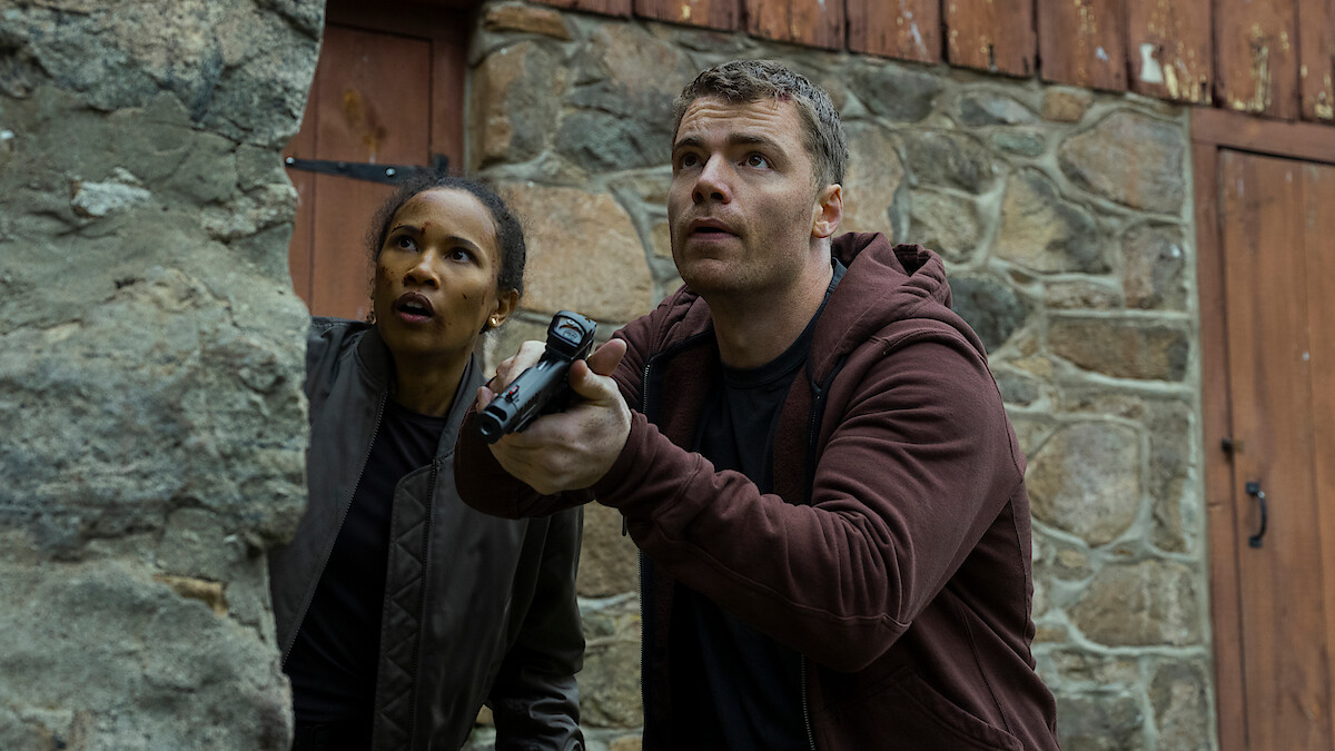 Two people crouch by a stone wall, looking alert and cautious. One holds a gun, both appear focused and ready for action. The setting is rustic, with stone and wood walls suggesting a rural or remote area.