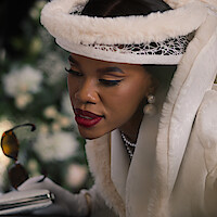 Woman in elegant white attire with pearls and fur hat looking into an open casket at a funeral, holding sunglasses and a clutch, with floral arrangements in the background.