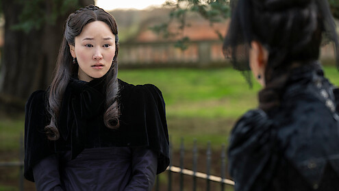 A woman in Victorian-style black clothing sits outdoors in a park or garden, looking concerned while talking to another person dressed similarly. Trees and a fence are visible in the background.