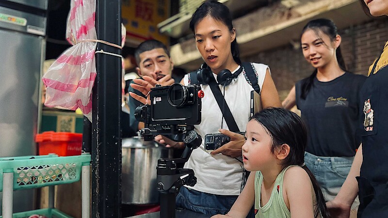 A woman shows a young girl and a group of people how to use a camera on a tripod in a market setting. The mood is focused and friendly, suggesting collaboration and learning in a lively outdoor environment.