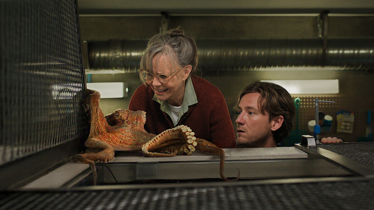 An older woman and a younger man observe an octopus on a metal surface in a laboratory or aquarium setting, with industrial pipes and equipment in the background.
