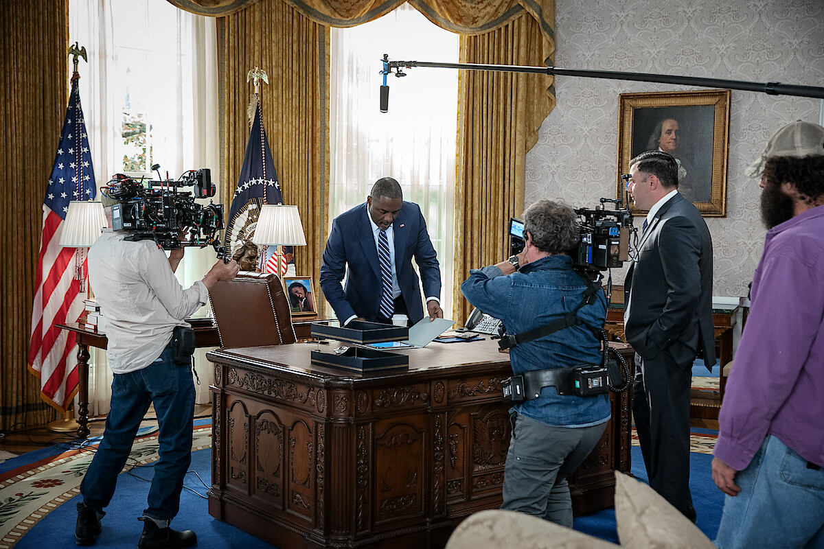 Actor in a suit stands at a presidential desk in an Oval Office set, surrounded by film crew, cameras, and lighting equipment, creating a busy, professional atmosphere for a political drama scene.