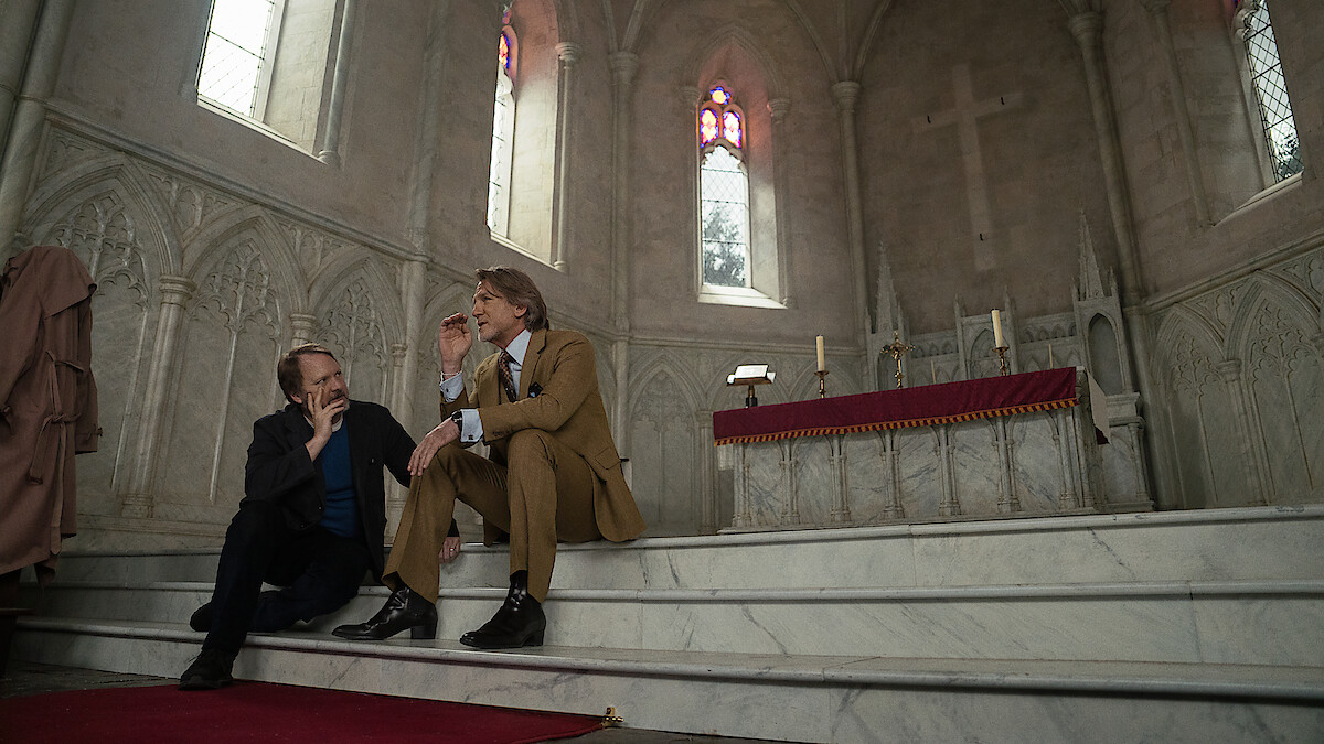 Two men in suits sit on marble steps in a gothic church, talking seriously near an altar with candles and stained glass windows; the setting feels solemn and contemplative.