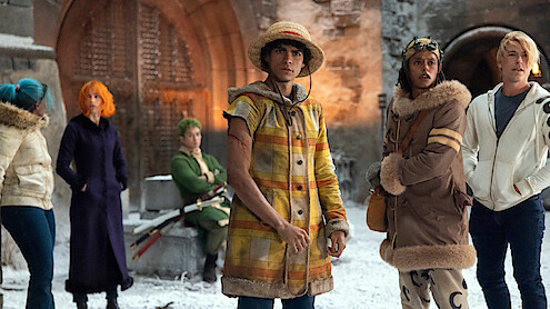 Six people in colorful, winter outfits stand in a snowy courtyard before a large gate, appearing alert or contemplative. The setting is medieval with stone walls and heavy wooden doors.