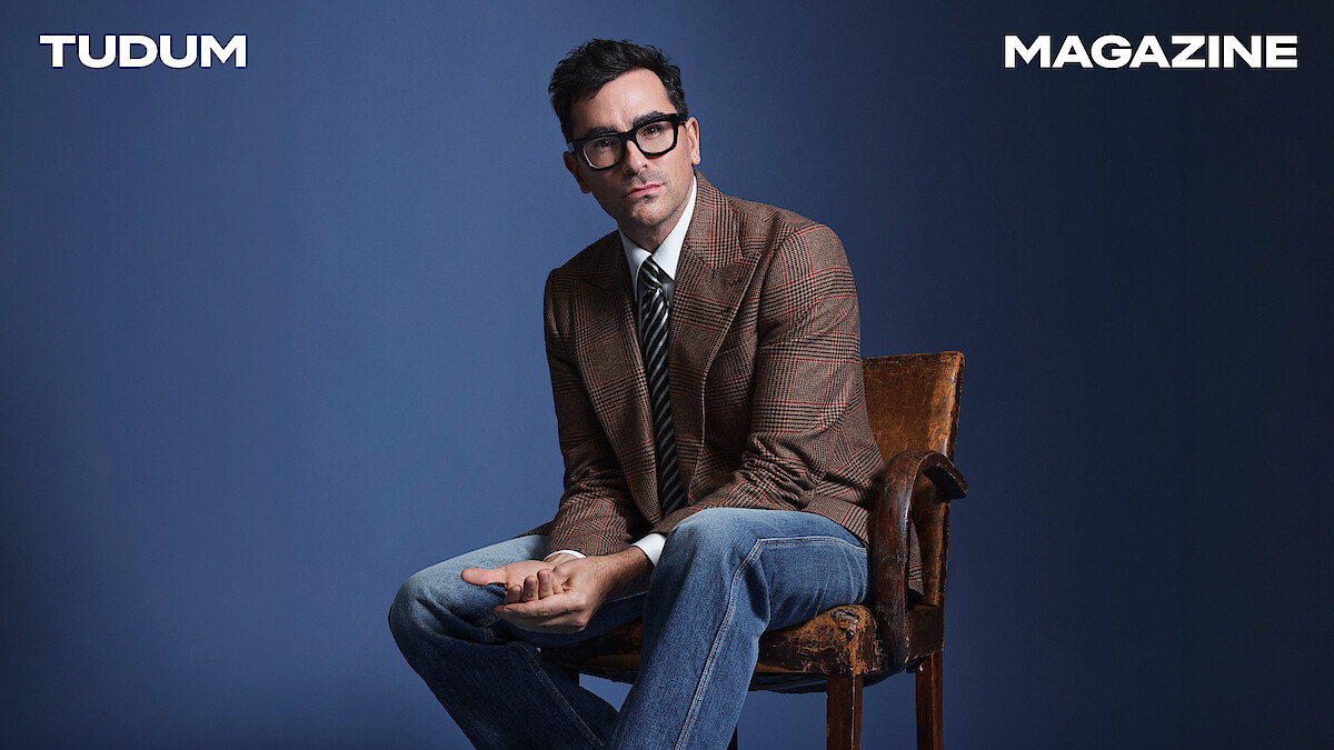 Dan Levy poses against a blue backdrop, wearing a checked blazer.