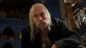 Elderly man with long white hair and mustache wearing dark robe, sitting indoors in a dimly lit room with blue and wooden details, serious and intense expression, medieval or fantasy setting, somber mood.