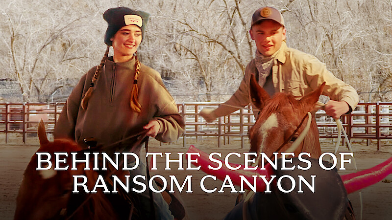 From left to right, Lizzy Greene and Garrett Wareing are smiling while riding horses in a fenced area with bare trees. Text reads "BEHIND THE SCENES OF RANSOM CANYON" at the bottom.
