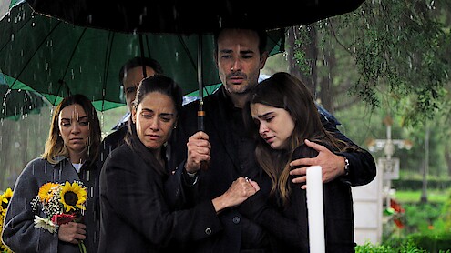 At a gravesite, actor Regina Blandón stands holding a bouquet of flowers behind Sebastián Martínez, playing Emiliano Lobo, who holds an umbrella over Ana Claudia Talancón, playing Daniela Lobo, as they embrace the their teenage daughter.