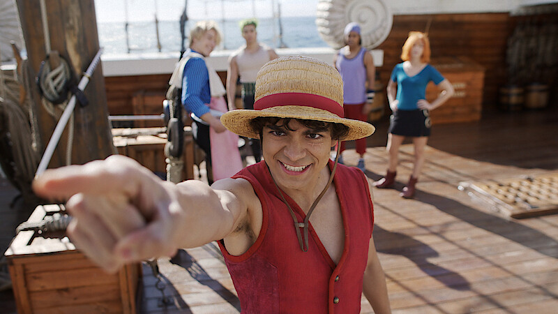Person in a straw hat and red vest pointing forward on a wooden ship deck, with ocean in the background and four people standing behind, dressed in colorful outfits, under bright daylight.