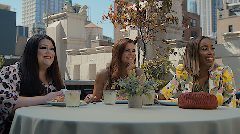 Three women sit at an outdoor rooftop table in a city, smiling and enjoying drinks and food, surrounded by buildings and greenery in a casual, urban daytime setting.
