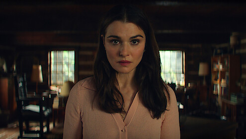 Rachel Weisz as M with long brown hair and a light pink blouse stands indoors, looking directly at the camera in a warmly lit, rustic room with wooden walls, windows, and cozy furnishings.
