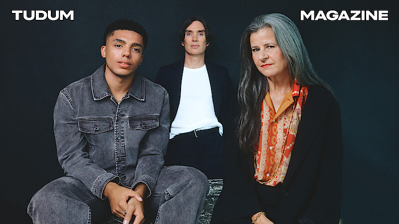 From left to right, Jay Lycurgo, Cillian Murphy, and Tracey Ullman sit around a table against a dark slate grey background.