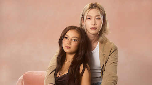 Two people sit closely together on a pink chair in a studio with a soft, pastel-colored background. One person wears glasses and a dark dress, the other has long blonde hair and a tan jacket, both looking at the camera.