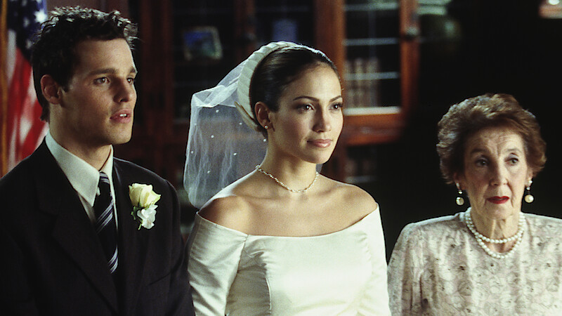 A bride in a white dress and veil stands between a man in a suit with a boutonniere and an elderly woman in pearls, indoors with bookshelves and an American flag in the background.