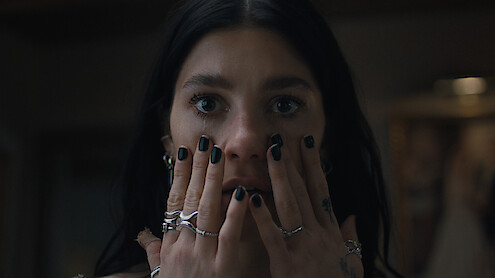A woman with dark hair and black nail polish holds her hands to her face indoors, wearing multiple rings and looking surprised or concerned in a dimly lit setting.