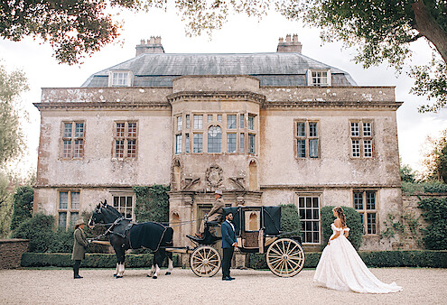 Bride in a white gown stands near a vintage horse-drawn carriage outside an elegant, historic mansion. Lush greenery frames the scene, evoking a romantic, fairytale atmosphere. The mood is dreamy and regal.
