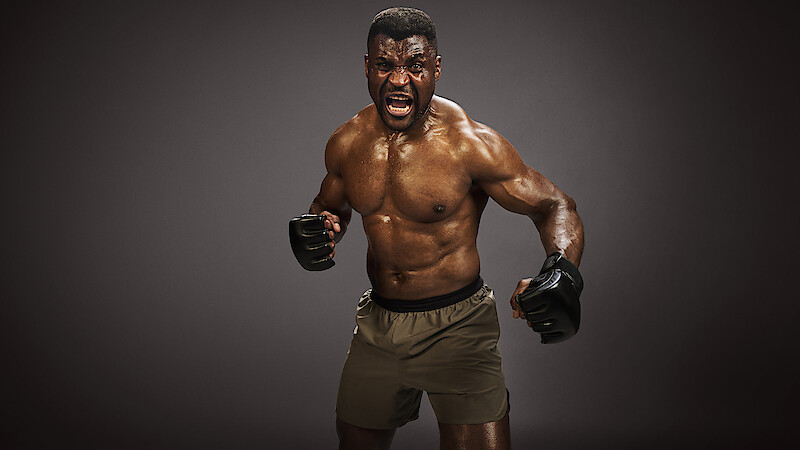 Francis Ngannou in a fighting stance, shirtless and wearing gloves and shorts, yelling intensely in a dimly lit studio with dark background.