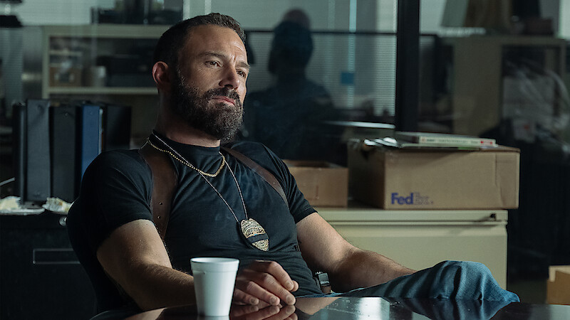 Bearded man wearing a badge and shoulder holster sits relaxed at a desk in an office, coffee cup in hand, with boxes and files in the background, giving a detective or law enforcement vibe.