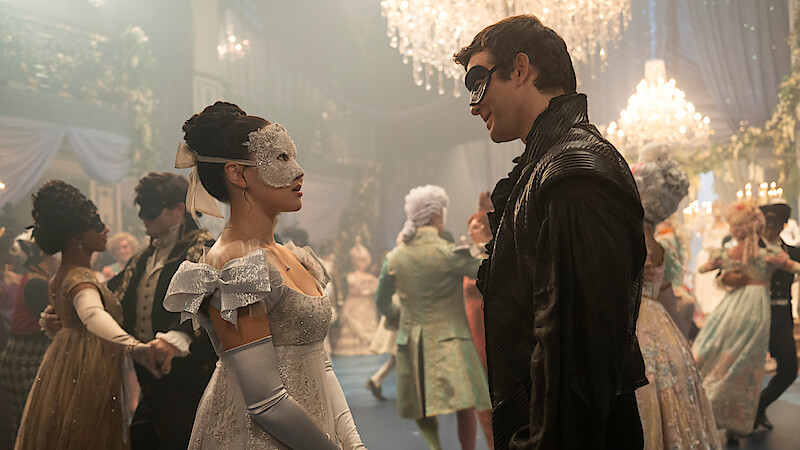 Two people in elegant masquerade costumes face each other at a grand ballroom, surrounded by guests in period attire and ornate chandeliers, evoking a formal and festive historic setting.