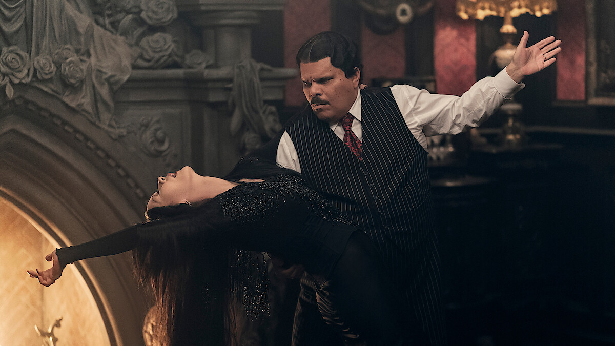 Catherine Zeta-Jones as Morticia Addams and Luis Guzmán as Gomez Addams dancing together in front of a fireplace in 'Wednesday' Season 2.