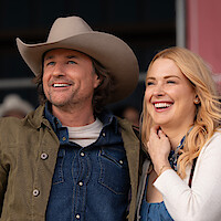 A smiling man in a cowboy hat and a woman with blonde hair stand close together outdoors, both looking happy and relaxed, suggesting a casual, friendly atmosphere.