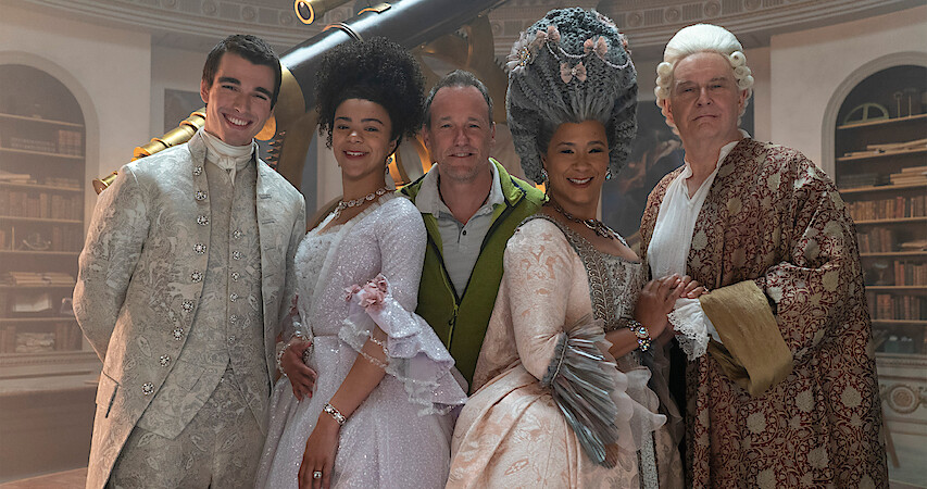 The young and older versions of Queen Charlotte and King George smile with director Tom Verica