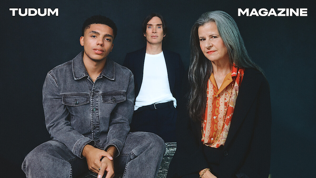 From left to right, Jay Lycurgo, Cillian Murphy, and Tracey Ullman sit around a table against a dark slate grey background.