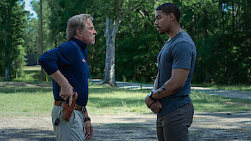 Two men stand outdoors facing each other in a grassy, wooded area. One man, older and wearing a blue shirt with khaki pants, has a hand on a holstered gun. The other, younger man, stands calmly with hands clasped.