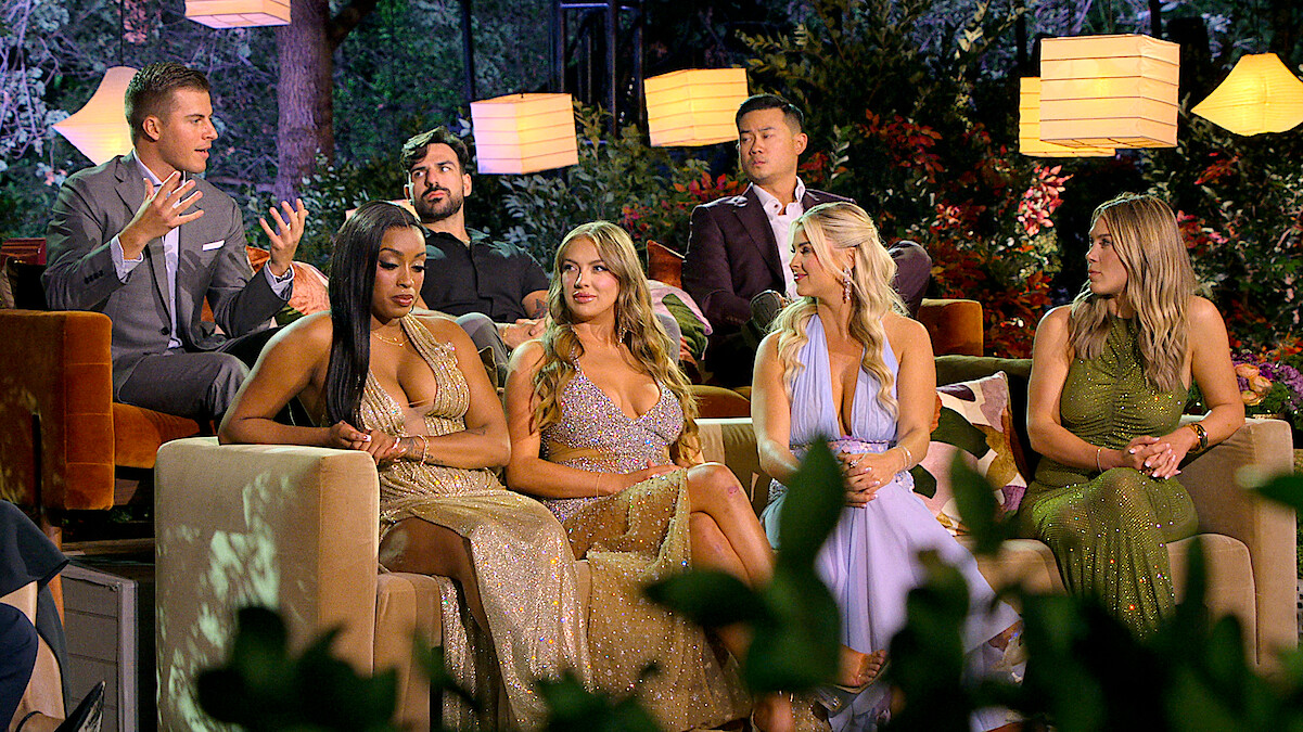 Six people in formal attire sit on couches outdoors at night, surrounded by lush greenery and hanging lamps, engaged in conversation with a vibrant, glamorous atmosphere.