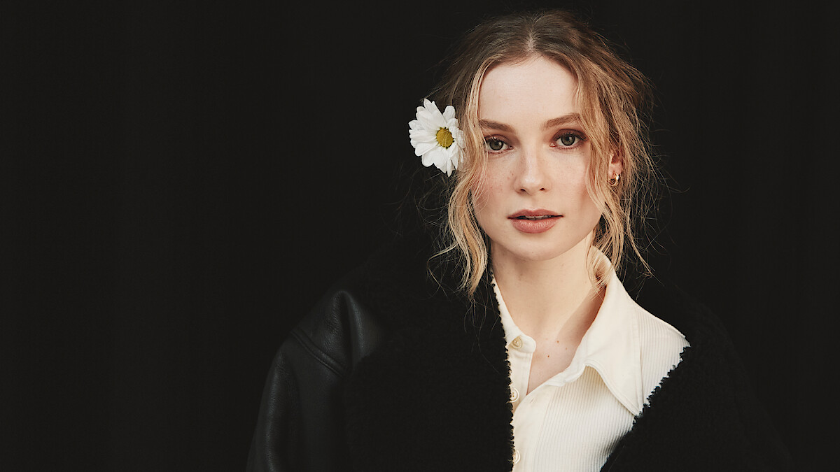 Woman with wavy blond hair and a white flower tucked behind her ear, wearing a white shirt and black coat, posed in front of a dark background.