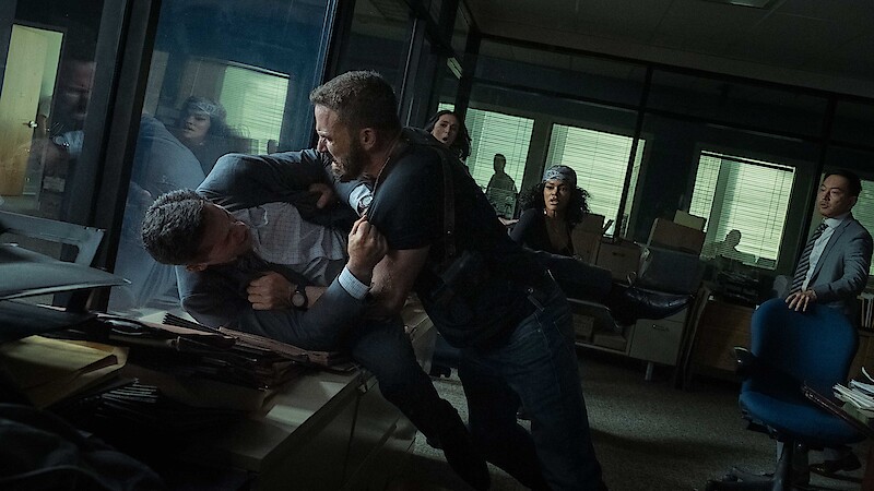 Two men struggle violently in an office, one pinning the other against a desk, while others watch in shock. The setting is a cluttered workspace with glass walls and scattered files, lit by daylight through window blinds.
