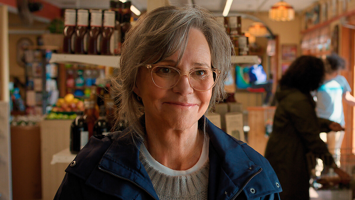 Smiling older woman with gray hair and glasses in a blue jacket stands inside a well-lit grocery store, with shelves of food and drinks, people shopping, and produce in the background.