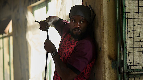A man in a red shirt and headwrap cautiously holds a rope while pressed against a wall inside a dimly lit building with barred windows, appearing alert and wary of his surroundings.